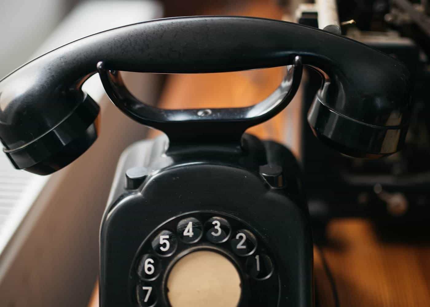 Black rotary telephone on a wooden table beside a typewriter