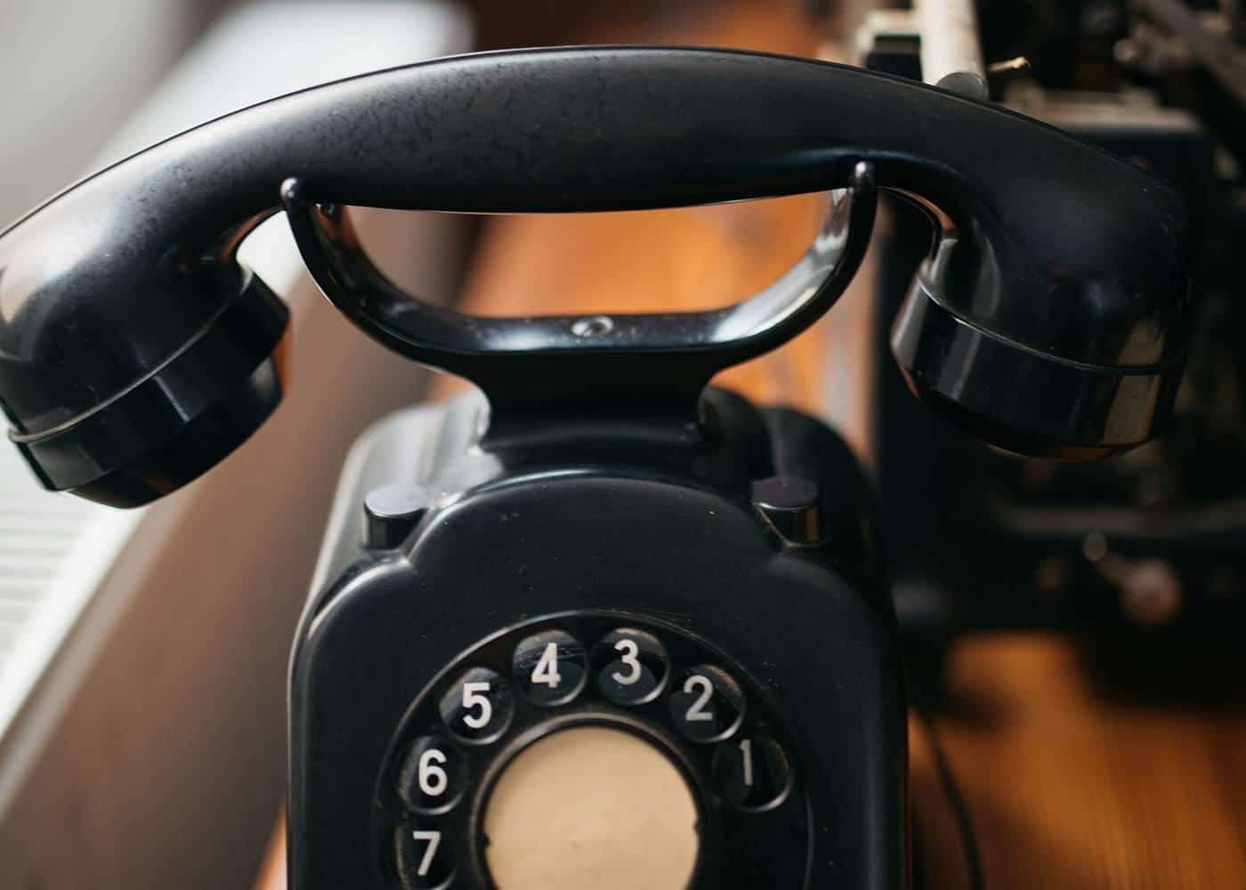 Black rotary telephone on a wooden table beside a typewriter
